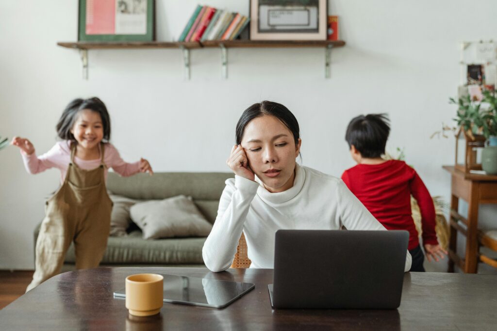 Stressed mother working on a laptop while children play in the background, representing high-functioning anxiety in mothers balancing work and family.